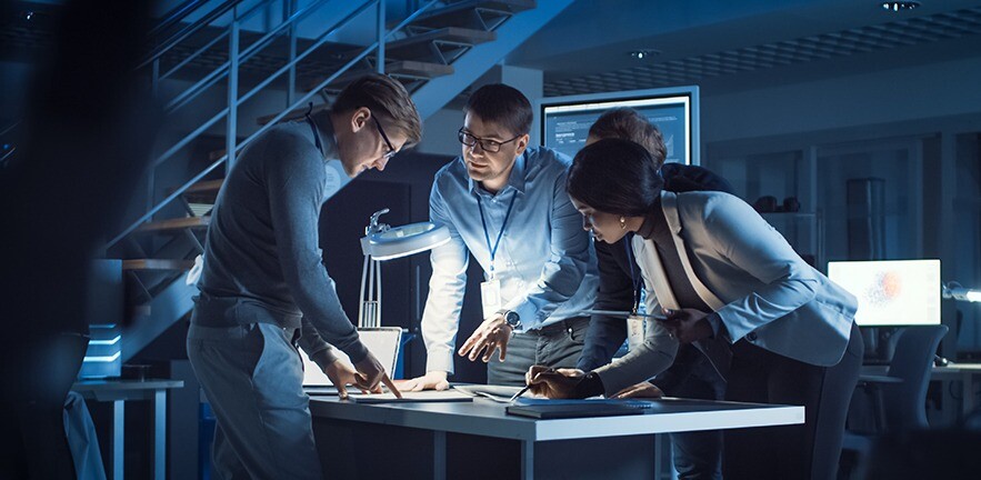 A diverse team of electronics development engineers standing around a desk late at night, solving a project problem.