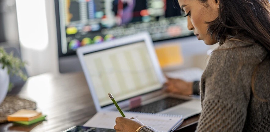 Woman tracking investments on a laptop, making notes on her notepad.