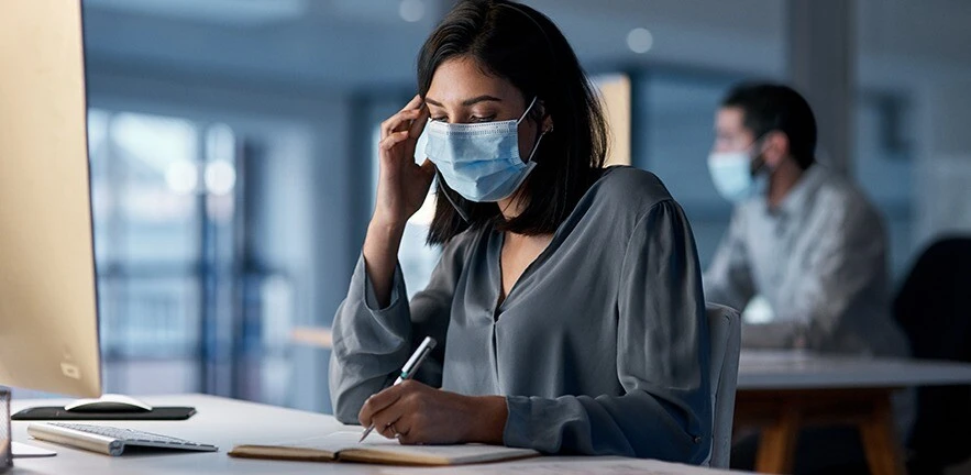 Shot of a woman using a headset and computer while wearing a mask in a modern office.