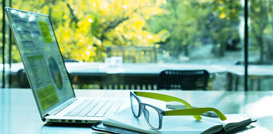 Laptop computer on a table with a pair of glasses on a notepad beside it in an open-plan office with sunny garden outside window.