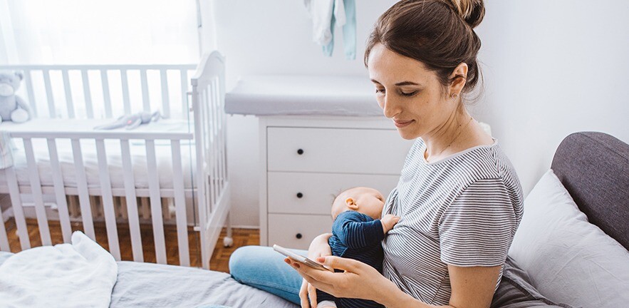 Mother breastfeeding her baby son in the bedroom while using a mobile phone.