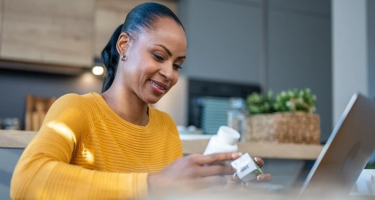 Smiling mature woman looking at pill bottle while using laptop in kitchen.